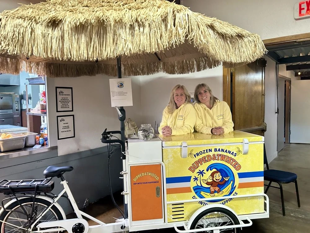 Two people behind the yellow Dipped & Twisted bicycle cart, with blue and orange stripes and a thatched parasol.
